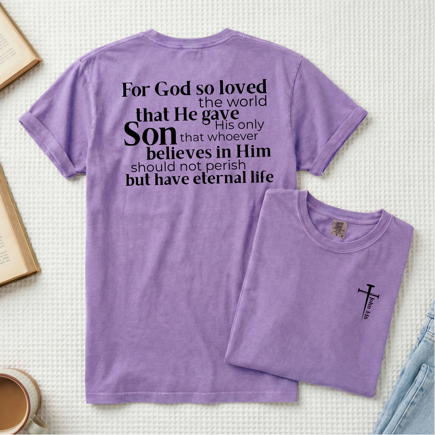 A purple short-sleeve T-shirt laid flat on a light textured background, with a smaller folded version placed in front. The main shirt shows a large black typographic religious quote on the back reading, 'For God so loved the world that He gave His only Son who whoever believes in Him should not perish but have eternal life.