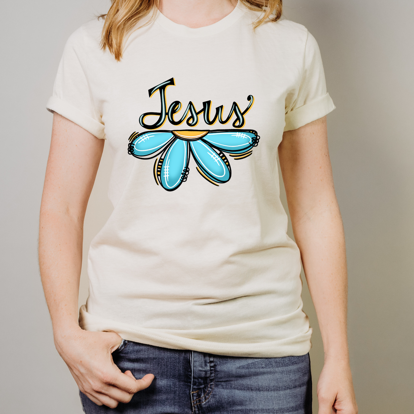 A person wearing a cream-colored t-shirt with a graphic that reads 'Jesus' in black script above a stylized light blue flower with gold outlines, paired with blue jeans.