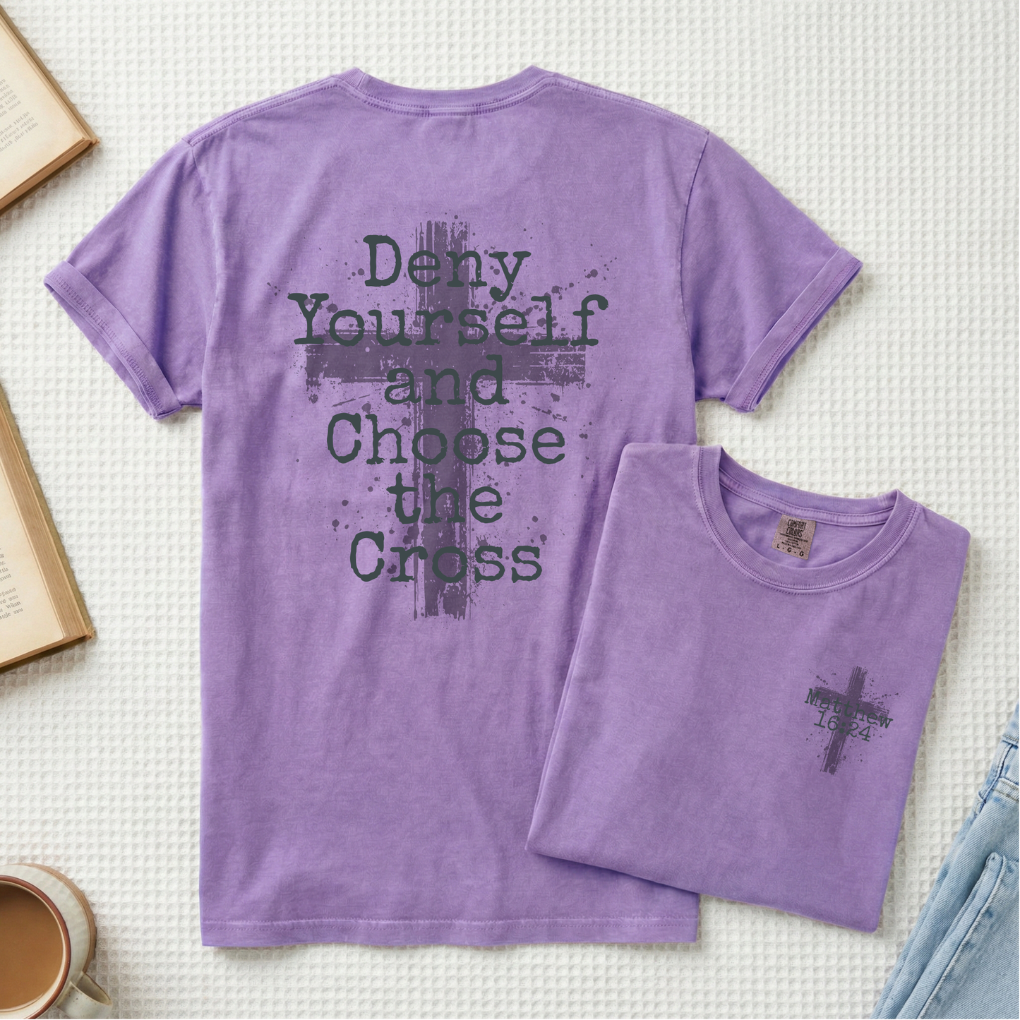 A purple short-sleeve T-shirt shown flat-laid on a light textured background, with a folded matching T-shirt placed in front. The larger shirt shows the back with a dark gray distressed cross and the text 'Deny Yourself and Choose the Cross.