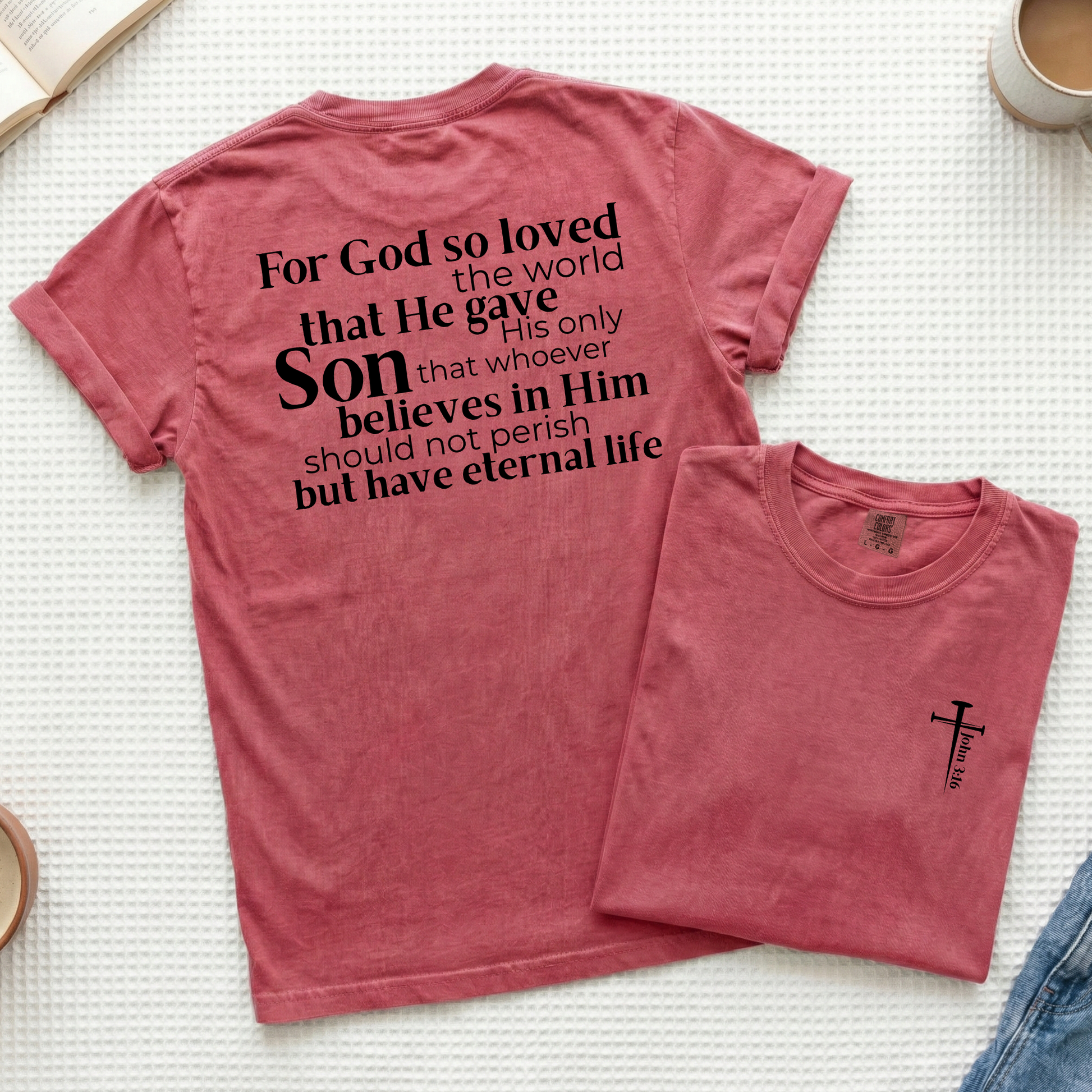 A dusty rose short-sleeve t-shirt shown flat-laid, with the back view displayed larger and a folded view of the same shirt placed in front. The back features a black typographic Christian message about God loving the world and giving His only Son.