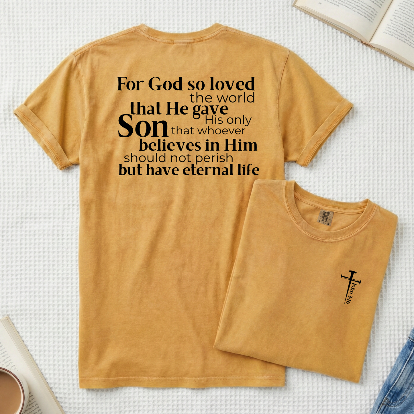 A mustard yellow t-shirt is laid flat on a white textured blanket, showing the back with black printed Bible verse text. A smaller folded view of the same shirt is placed in front, showing a small black cross and 'Jesus Loves Me' print.