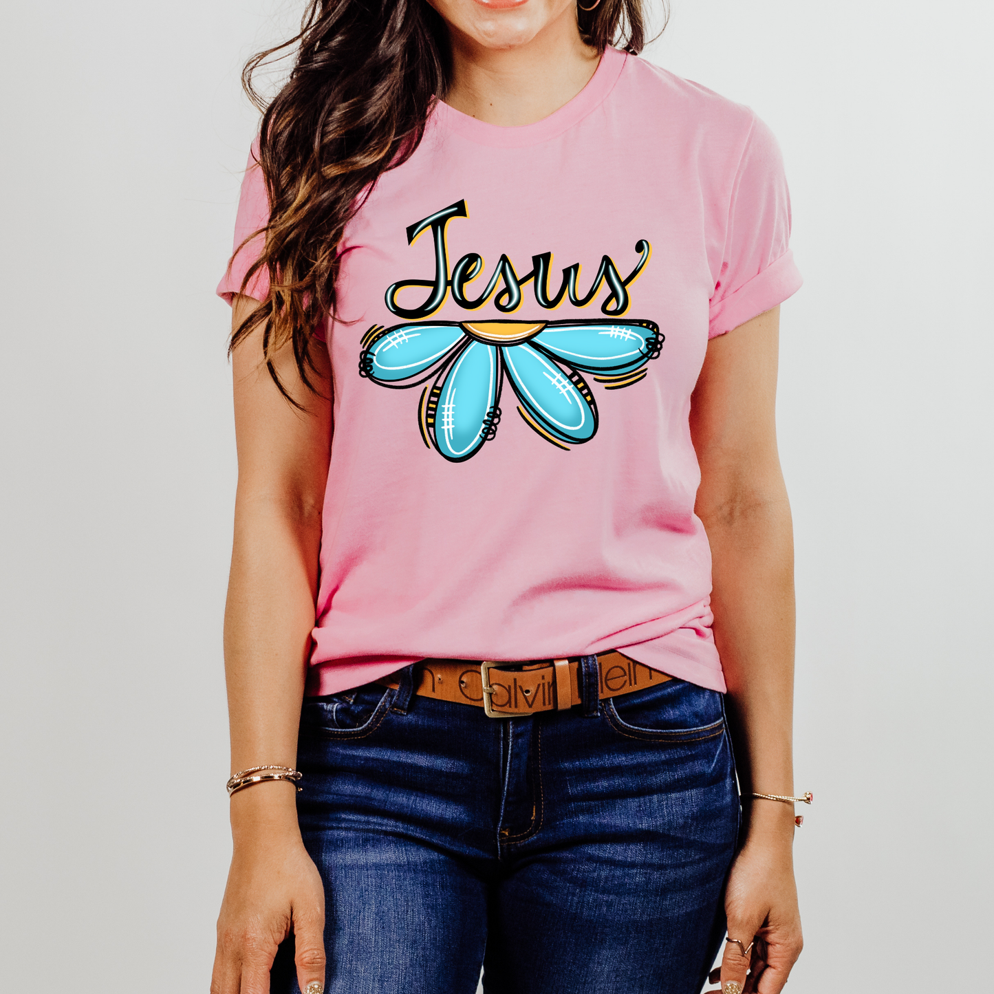 A woman wearing a pink short-sleeve graphic t-shirt with 'Jesus' written in black script above a light blue flower, paired with dark blue jeans and a brown belt.