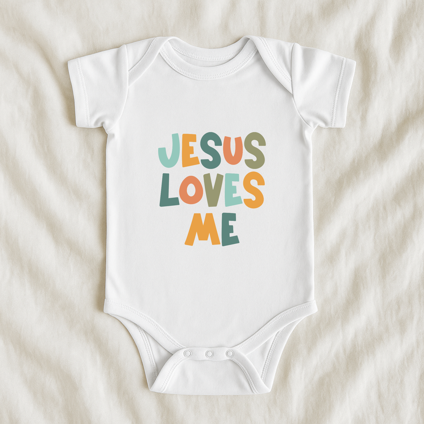 A white short-sleeve baby bodysuit laid flat on a soft beige fabric background, featuring the phrase 'JESUS LOVES ME' printed in colorful retro-style letters.