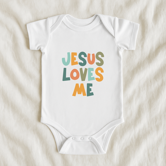 A white short-sleeve baby bodysuit laid flat on a soft beige fabric background, featuring the phrase 'JESUS LOVES ME' printed in colorful retro-style letters.