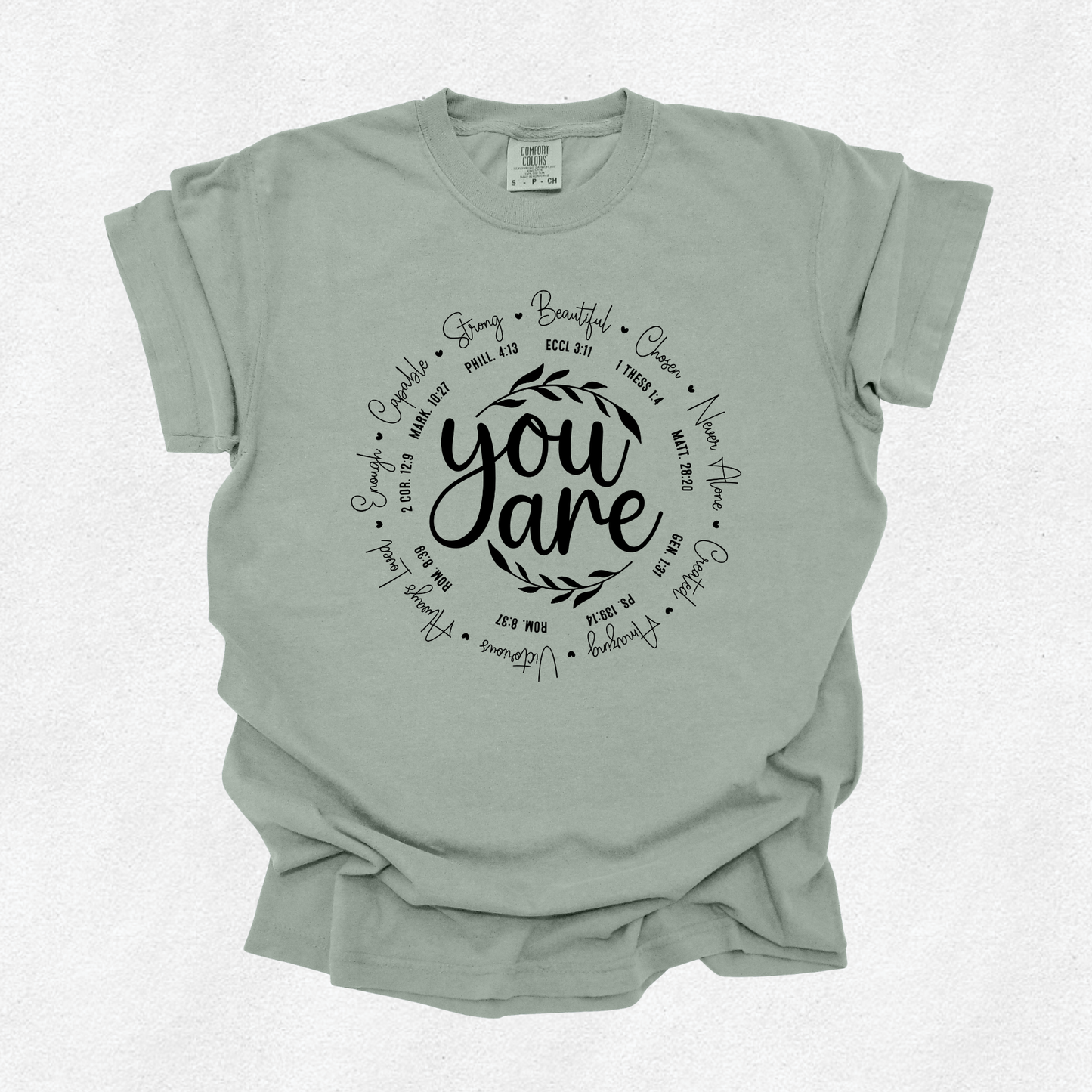 A sage green short-sleeve t-shirt with a black graphic print on the front reading 'you are' surrounded by inspirational words, laid flat on a light textured background.