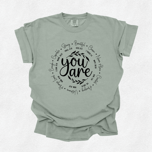 A sage green short-sleeve t-shirt with a black graphic print on the front reading 'you are' surrounded by inspirational words, laid flat on a light textured background.