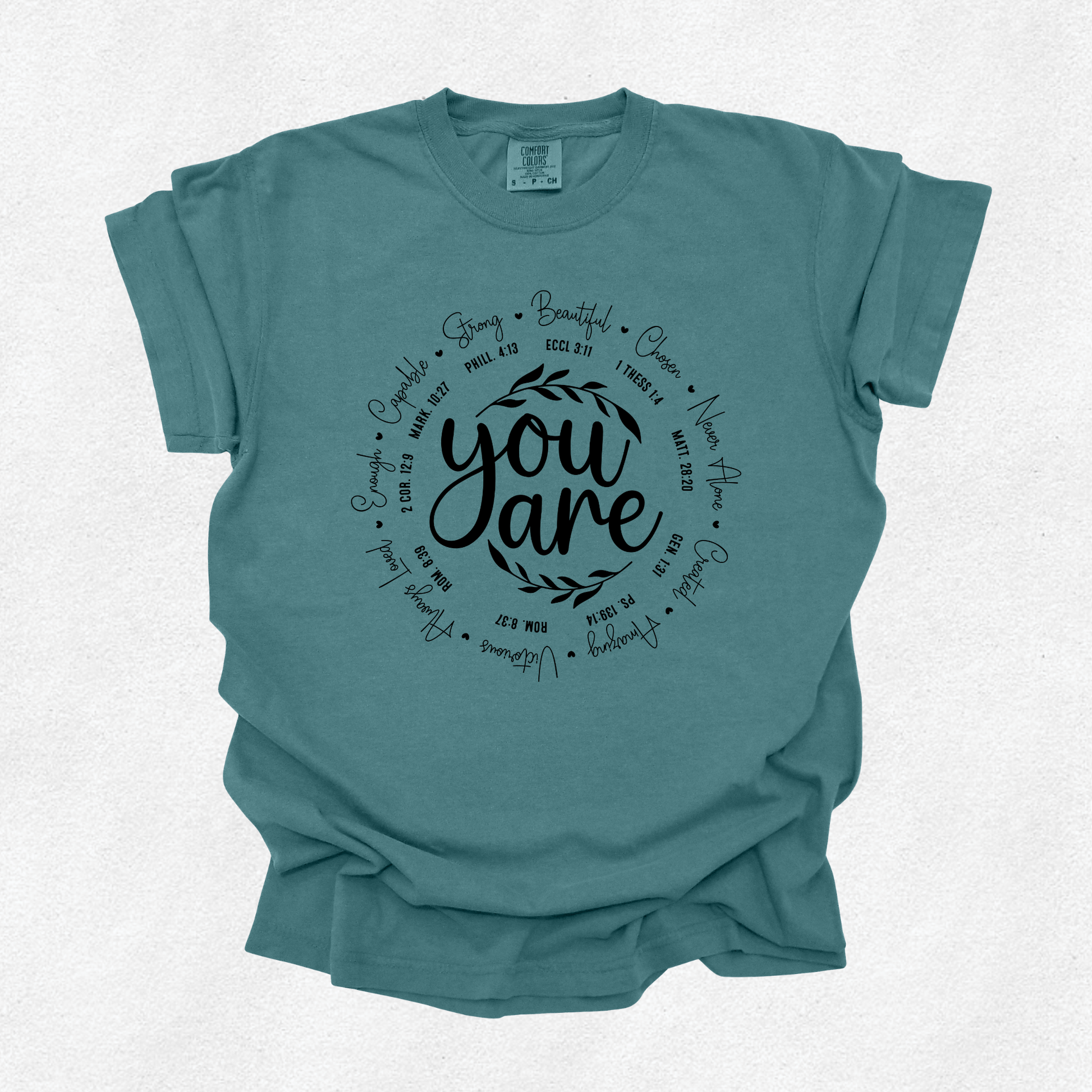 A teal blue short-sleeve t-shirt with a black graphic print on the front. The graphic features the words 'you are' in a central script font, surrounded by a circular arrangement of various biblical scripture verses.