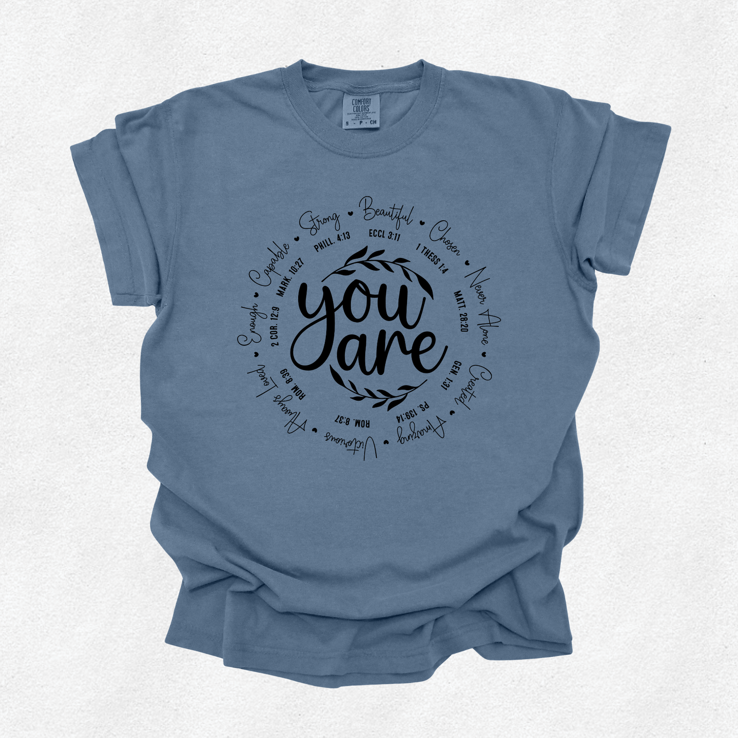 A dusty blue short-sleeve t-shirt with a black graphic print on the front reading 'you are' surrounded by a circle of inspirational adjectives, laid flat on a light textured background.