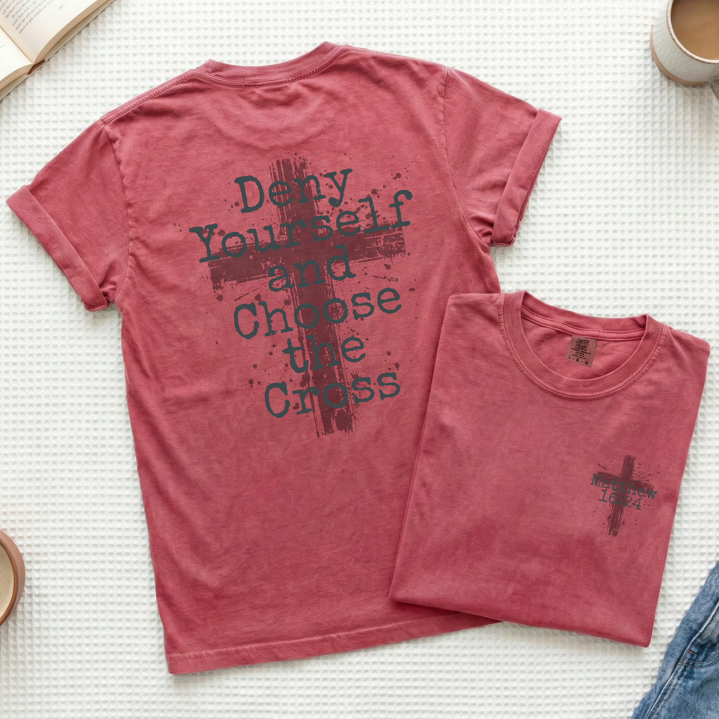 A dusty rose short-sleeve t-shirt shown flat-laid, with the back view featured prominently and a smaller folded view placed in front. The back has a dark distressed cross graphic and the text 'Deny Yourself and Choose the Cross.