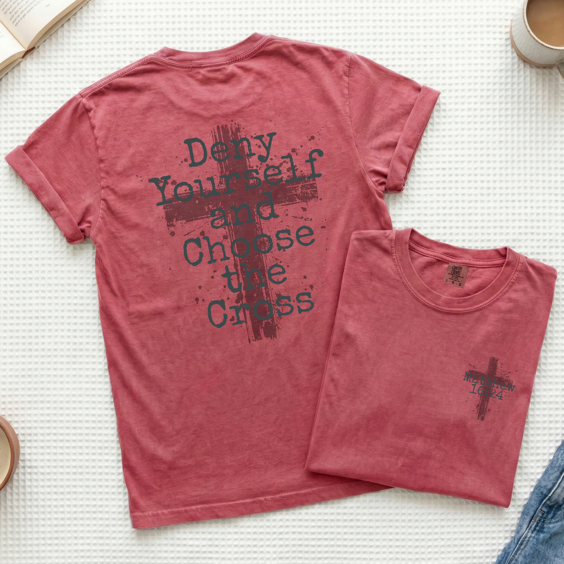 A dusty rose short-sleeve t-shirt shown flat-laid, with the back view featured prominently and a smaller folded view placed in front. The back has a dark distressed cross graphic and the text 'Deny Yourself and Choose the Cross.