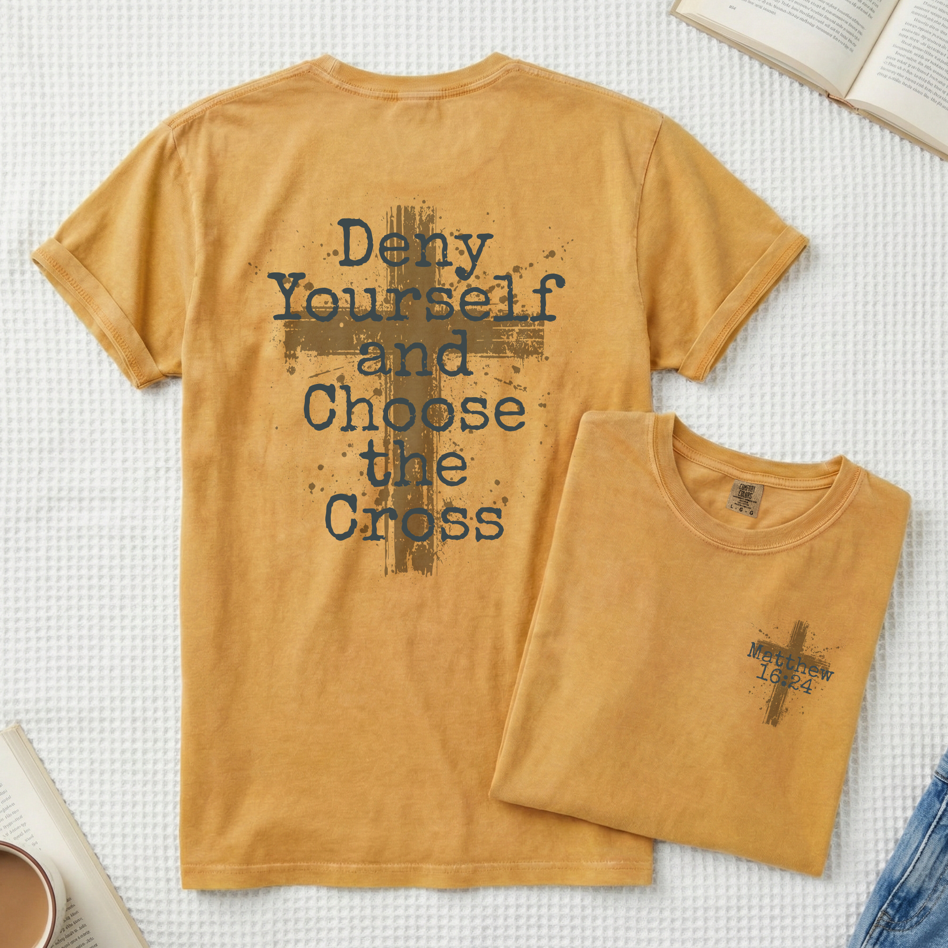 A mustard yellow t-shirt shown flat-laid on a white textured blanket, displaying the back with a dark blue typographic design that reads 'Deny Yourself and Choose the Cross' over a distressed cross. A smaller folded view of the front is shown next to it, featuring a small matching cross graphic with 'Fellowship of the Cross' text.