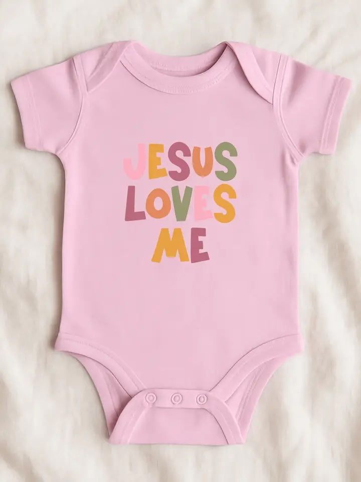 A light pink short-sleeve baby bodysuit laid flat on a soft white textured fabric background. The front features the phrase 'JESUS LOVES ME' in a playful, multicolored hand-drawn font.